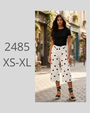 Mujer de pie en un callejón luciendo camiseta negra y pantalones blancos con puntos negros.