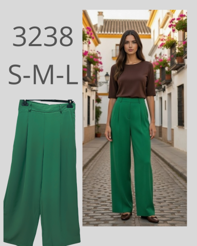 Mujer de pie en una calle con flores, vistiendo pantalones verdes y camiseta marrón.