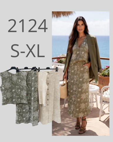 Mujer luciendo vestido largo con estampado floral frente al mar, rodeada de ropa en perchas.