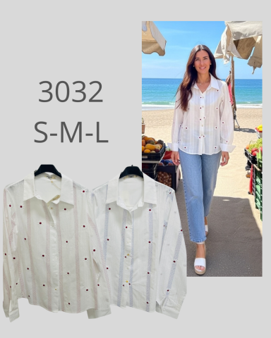 Mujer sonriente con camisa blanca de puntos, de pie en una playa.