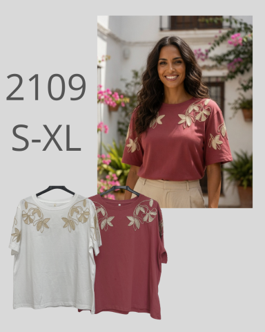 Mujer sonriendo con camiseta bordada y fondo de flores en un entorno exterior.