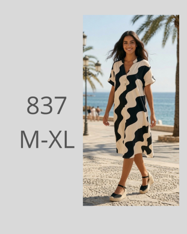 Mujer sonriente con vestido negro y blanco caminando junto al mar en un ambiente soleado.