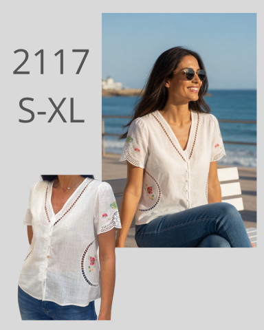 Mujer sonriente con blusa blanca y bordados, sentada junto al mar.