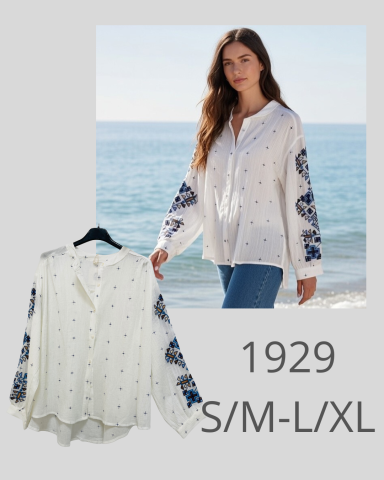 Mujer de pie junto a la playa, vistiendo una blusa blanca con detalles decorativos.