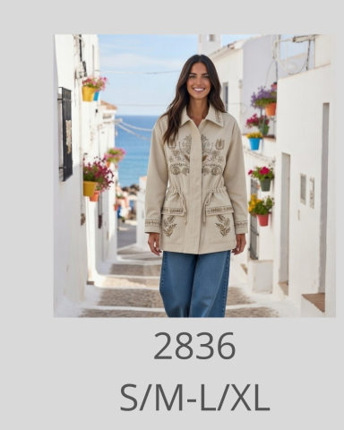 Modelo de abrigo beige con bordados, caminando por un pintoresco paisaje costero.