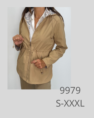 Mujer con chaqueta beige y blusa blanca, de pie con la mano en la cinta de la chaqueta.