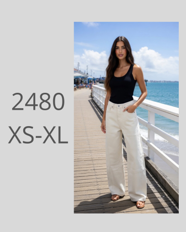 Mujer de pie en un paseo marítimo, usando top negro y pantalones claros, con mar de fondo.