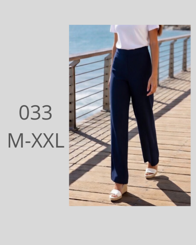 Mujer caminando en pantalones navy, con un fondo de playa y barandilla de madera.