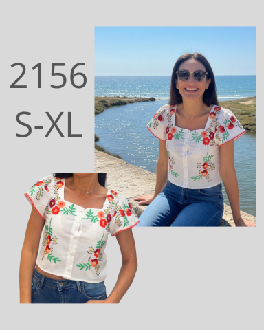 Mujer sonriente con blusa bordada de flores, sentada junto al agua.
