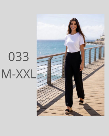 Mujer con camiseta blanca y pantalones negros, sonriendo en un muelle junto al mar.