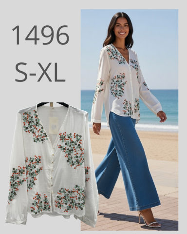 Mujer caminando con blusa blanca con flores y pantalones anchos, junto a la playa.