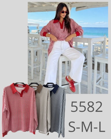 Mujer sentada con camisa de cuadros y pantalones blancos, fondo de playa y varias camisetas.