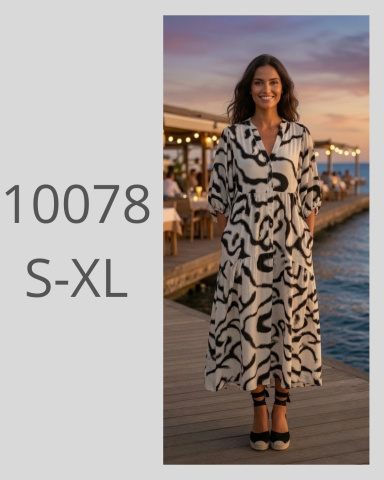 Mujer sonriente con vestido blanco y negro, de pie junto al agua al atardecer.