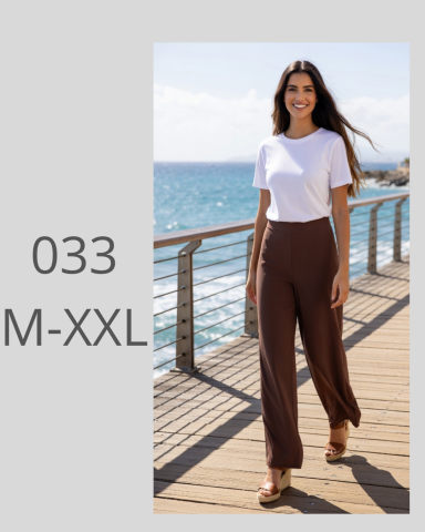 Mujer sonriente con camiseta blanca y pantalones marrones, caminando junto al mar.